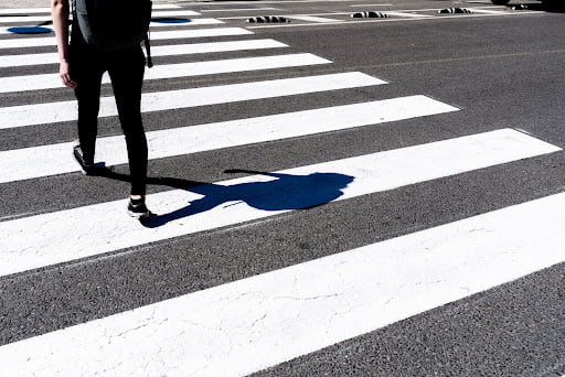 A person walks across a pedestrian crosswalk on a sunny day, casting a shadow on the asphalt.